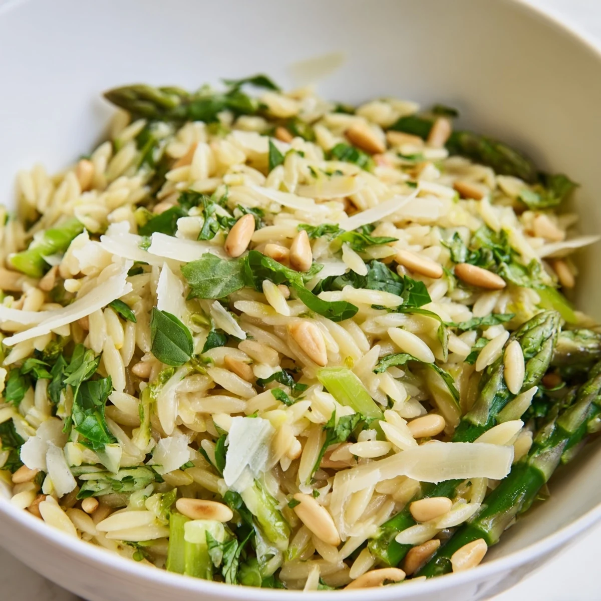 A close-up view of a vibrant Asparagus Lemon Orzo Salad served in a white bowl, garnished with toasted pine nuts and fresh parsley.