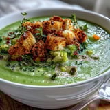 Spoon diving into a velvety puree of cauliflower and broccoli soup garnished with crunchy, oven-baked croutons.